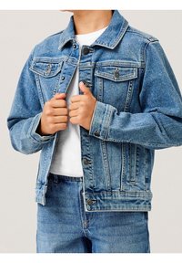 Džínová bunda světle modré barvy s dvěma kapsami na hrudi, kovovými knoflíky a klasickým límcem. Spárováno s odpovídajícími džínovými kalhotami a bílou košilí.