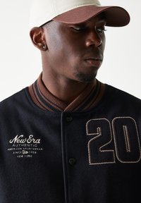 Μπλε ναυτικό μάλλινο varsity jacket με ριγωτό καφέ ριμπ λαιμόκοψη, που διαθέτει το λογότυπο "New Era" και μεγάλο κεντημένο "20" σε μπεζ ραφή.