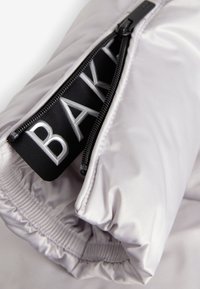 Puffige weiße Jackenstoff mit einem Reißverschluss, der eine schwarze Schlaufe mit dem geprägten Schriftzug „BAK“ aufweist. Glatte Textur und elastischer Saum.