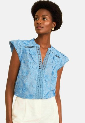 Vrouw draagt een lichtblauwe mouwloze blouse met bloemenborduursel en kanten afwerking en crème kleurige broek, kijkt opzij.
