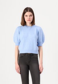 Maglione azzurro chiaro a maniche corte con finitura testurizzata e maniche a sbuffo, abbinato a jeans scuri. Collo rotondo e dettagli a coste sul bordo.