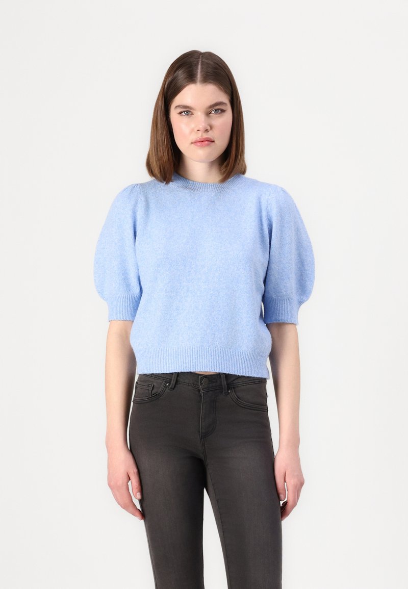 Maglione azzurro chiaro a maniche corte con finitura testurizzata e maniche a sbuffo, abbinato a jeans scuri. Collo rotondo e dettagli a coste sul bordo.