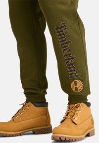 Grüne Sweatpants mit "Timberland"-Logo und Baum-Grafik. Beige Stiefel mit einer strukturierten Wildleder-Oberfläche, Metallösen und Gummisohlen.