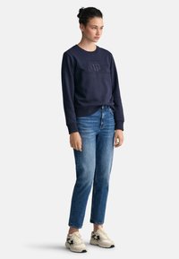 Felpa in cotone navy con logo in rilievo, abbinata a jeans azzurri e sneakers beige con dettagli neri. Stile casual, vestibilità rilassata.