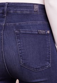 Jeans de mezclilla azul oscuro con un diseño ajustado, que cuentan con un bolsillo trasero, una etiqueta de marca y detalles de costura sutiles.
