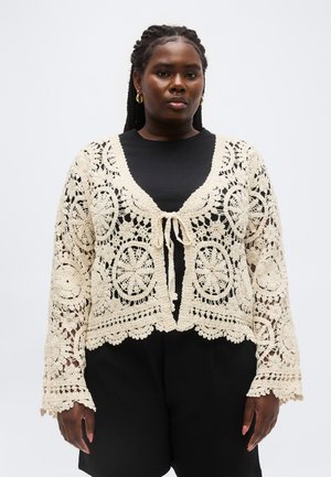 Frau trägt ein schwarzes Oberteil und Shorts sowie eine cremefarbene, offene Häkelstrickjacke, die vorne an der Brust gebunden ist, steht vor einem einfachen Hintergrund.