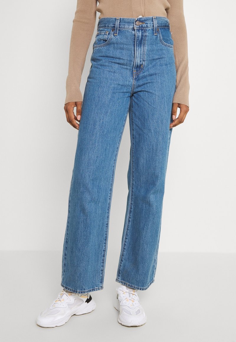 Personne portant un jean bleu taille haute, droit, un haut beige à manches longues et des baskets blanches épaisses, debout devant un fond blanc.