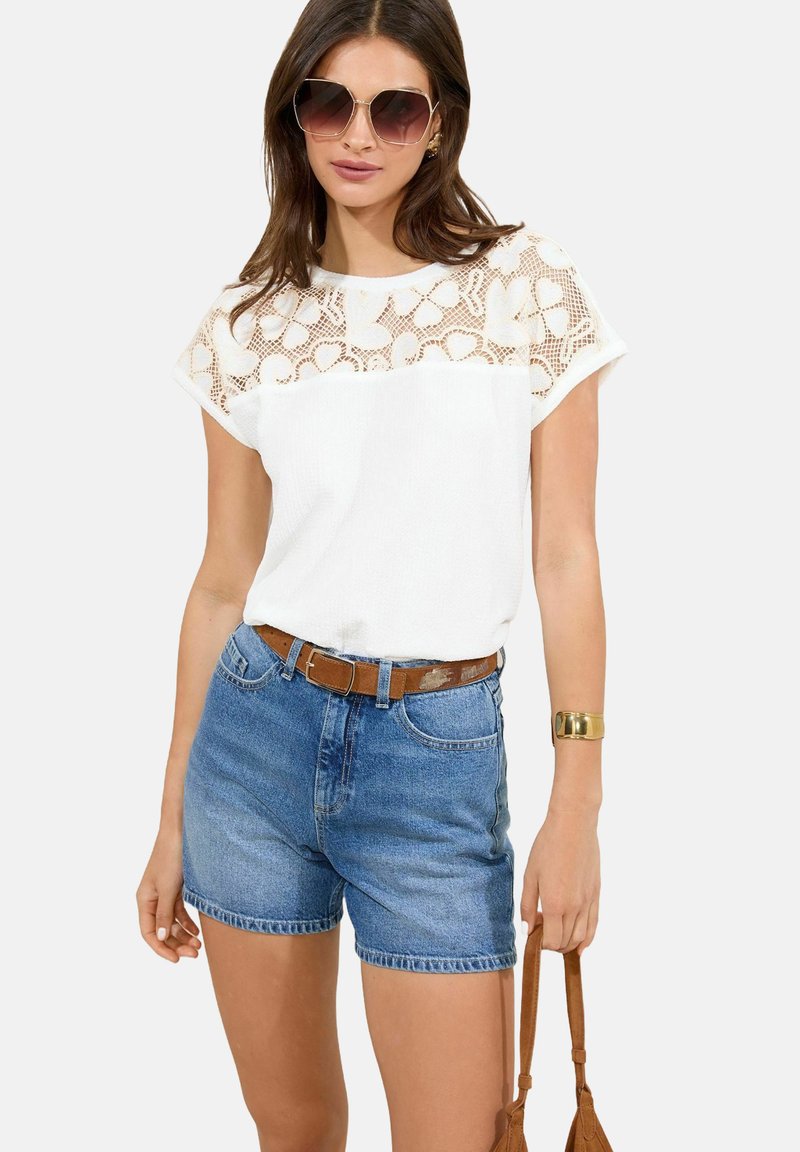 Haut blanc à manches courtes avec un motif en dentelle texturé aux épaules, associé à un short en denim bleu taille haute et une ceinture marron.