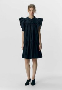 Schwarzes, gerafftes Kurzarmkleid mit einem gathered Taillenbereich, schichtweisem Design und geschwungenen Akzenten. Getragen mit schwarzen High Heels.