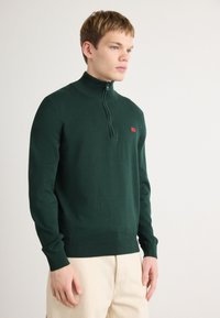 Dunkelgrüner Reißverschluss-Pullover mit hohem Kragen, gerippten Bündchen und Saum. Verfügt über ein kleines rotes Logo auf der Brust. Weicher Strickstoff.