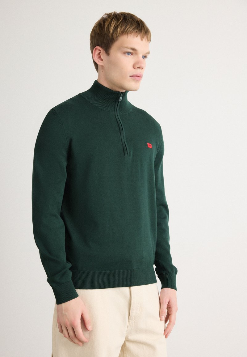 Dunkelgrüner Reißverschluss-Pullover mit hohem Kragen, gerippten Bündchen und Saum. Verfügt über ein kleines rotes Logo auf der Brust. Weicher Strickstoff.