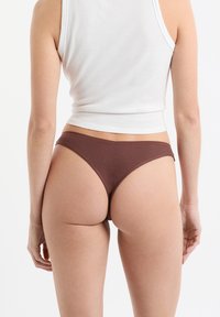 Perizoma marrone a coste con vita bassa e copertura posteriore minimalista, abbinato a un top bianco aderente senza maniche.