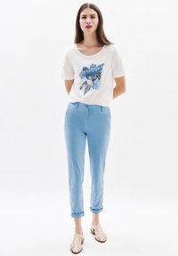 Weißes T-Shirt mit blauem Blumenmuster, kombiniert mit hellblauen Hosen in schmaler Passform, umgeschlagenen Säumen und beigen flachen Sandalen mit Riemchen.