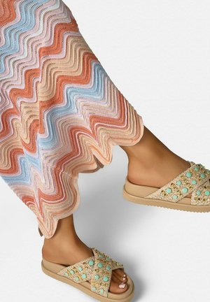 Pies con sandalias beige tipo slip-on con tachuelas turquesas, combinados con una falda o vestido de punto con patrón ondulado multicolor.