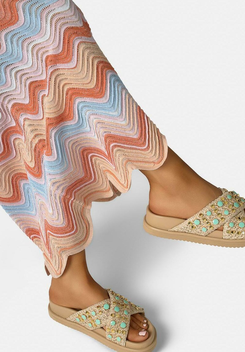 Füße in beigen Slip-On-Sandalen mit türkisfarbenen Nieten, kombiniert mit einem mehrfarbigen, wellenförmigen Strickrock oder Kleid.
