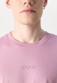 Růžové tričko s kulatým výstřihem vyrobené z měkké tkaniny s vyvýšeným logem "BOSS" ve stejném odstínu na přední straně. Detailní záběr na výstřih a texturu.