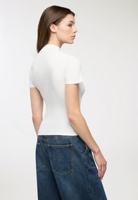 Donna con capelli castani lisci che indossa una maglietta bianca aderente a maniche corte e jeans di denim blu larghi, in vista di lato su uno sfondo semplice.