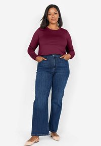 Haut à manches longues bourgogne avec des détails en dentelle, associé à un jean flare bleu en taille haute. Modèle portant des chaussures beiges, les mains dans les poches.