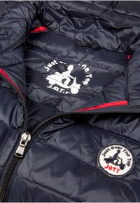 Interno di una giacca imbottita blu navy con accenti rossi. Presenta una zip argento, una texture trapuntata e due loghi: uno stampato e uno ricamato.