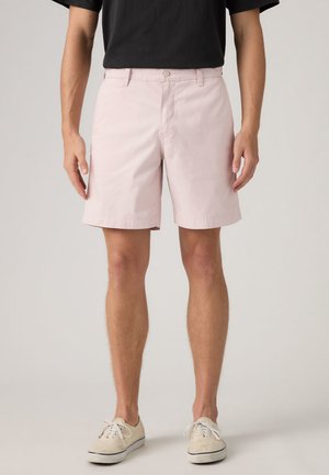 Uomo con pantaloncini rosa chiaro, t-shirt nera e sneakers beige basse, in piedi con le braccia rilassate lungo i fianchi.