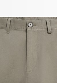 Gros plan sur la ceinture d'un pantalon beige avec un bouton en métal, des passants de ceinture et une fermeture éclair cousue.