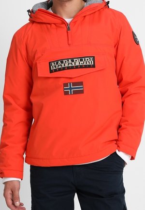 Homme portant une veste Napapijri orange vif avec poche à rabat avant, écusson du drapeau norvégien, et pantalon noir.