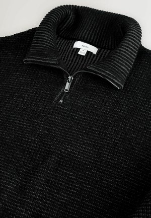 Svart zip-up-tröja med texturerad yta och ribbad krage. Har en central dragkedja och en etikett synlig inuti kragen.