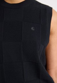 Svart ermeløs strikket topp med et teksturert sjakkmønster og en liten logo, laget av et mykt, elastisk materiale.