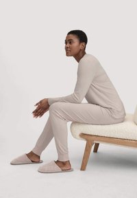 Hellbeige Loungewear-Set mit langen Ärmeln und eng zulaufenden Hosen, kombiniert mit sanft rosa Hausschuhen. Das Modell sitzt auf einer strukturierten, hellen Bank.