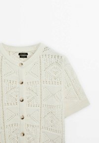 Knit cardigan in cream, featuring geometric cut-out patterns, short sleeves, and five large buttons down the front.
