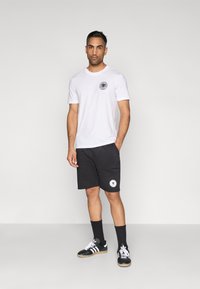 Λευκό κοντομάνικο μπλουζάκι με στρογγυλό μαύρο λογότυπο στο στήθος, σε συνδυασμό με σκούρο γκρι σορτς που έχει στρογγυλή επιγραφή, και μαύρα αθλητικά παπούτσια.