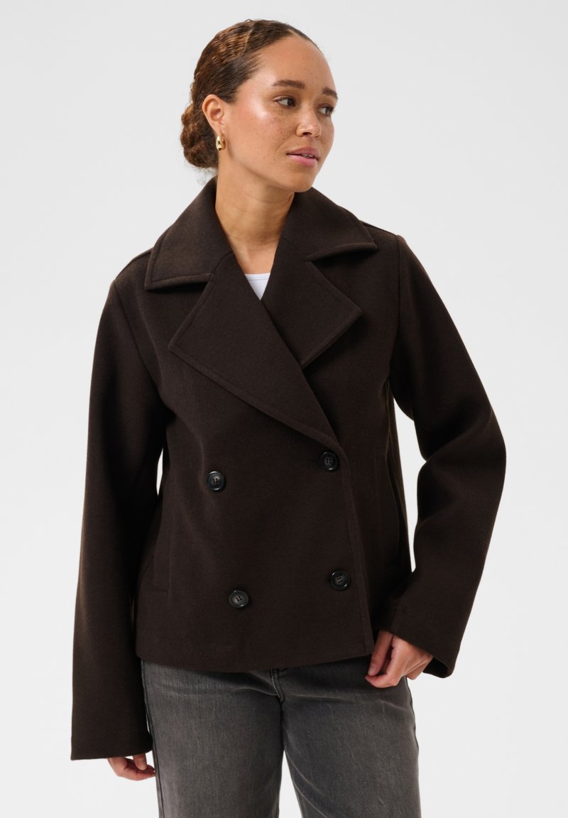 Femme portant un manteau en laine marron foncé à double boutonnage avec de larges revers, associé à un jean gris sur un fond clair uni.
