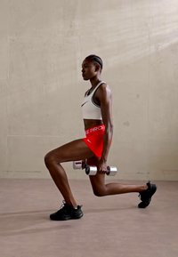 Athlete in a white sports bra and red Nike shorts, performing a lunge, holding dumbbells, wearing black athletic shoes on a light floor.