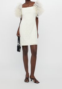 Weißes Spitzen-Minikleid mit quadratischem Ausschnitt und durchsichtigen, hellgrauen Tüll-Puffärmeln. Eine schwarze Handtasche und schwarze Riemchensandalen runden den Look ab.