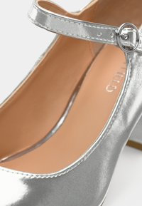 Zapato de salón metálico plateado con punta redonda, correa al tobillo con un hebilla circular y forro interior beige.