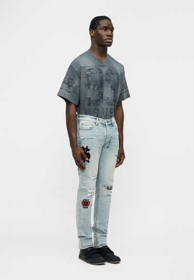 Άνδρας που στέκεται φορώντας μια γκρι φαρδιά μπλούζα με τον αριθμό 99, ανοιχτόχρωμα τζιν με φθορές και μπαλώματα, και μαύρα αθλητικά παπούτσια, μπροστά σε απλό φόντο.