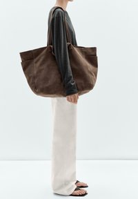 Brun mockaläder tote-väska med en bred, mjuk design och två ytterfickor, bärs över axeln, ihop med ljusa pants.
