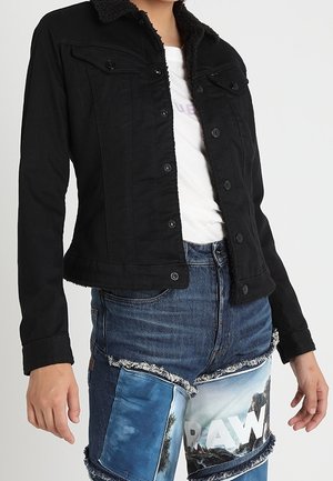Personne portant une veste noire boutonnée sur un t-shirt blanc et un short en jean bleu avec des détails graphiques et des patchs.