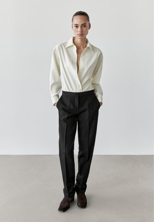 Kvinde stående med hænderne i lommerne, iført cremefarvet skjorte med knapper, sorte skræddersyede bukser og mørkebrune sko mod en ensfarvet baggrund.