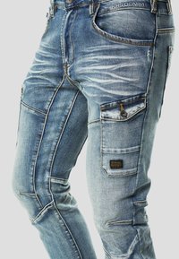 Světle modré džíny typu cargo z denimu s opranými detaily, několika kapsami, tmavým šitím a tkanou etiketou na boční kapse.