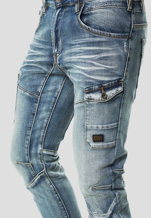 Világoskék farmer cargo nadrág, amely szakadt részletekkel, több zsebbel, sötét varrással és egy szövött címkével az oldalzseben.