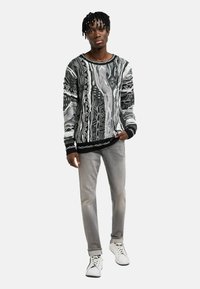 Schwarz-weiß gemusterter Pullover mit langen Ärmeln, kombiniert mit hellgrauen Jeans und weißen Sneakern. Der Pullover hat einen gerippten schwarzen Saum.