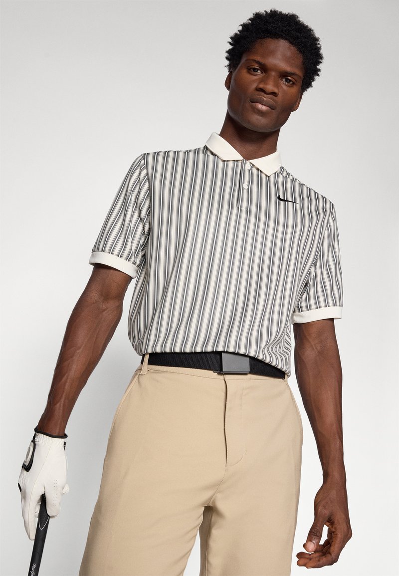 Polo a righe con colletto bianco e maniche corte, abbinato a pantaloni beige. La maglietta presenta righe verticali in grigio e nero.