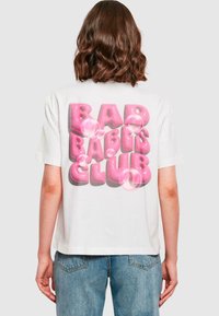 Hvit t-skjorte med korte ermer som har en stor grafikk i rosa bobletype på baksiden med teksten "BAD BABIES CLUB," med transparente boble-detaljer.