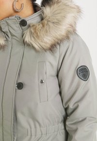 Femme portant un manteau d'hiver gris clair avec capuche bordée de fourrure et patch noir sur la manche, avec une boucle d’oreille anneau étincelante visible.
