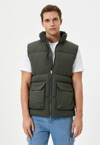 Chaleco acolchado verde oscuro con capucha, cremallera frontal y dos bolsillos en el pecho. Material texturizado con un acabado suave y un ajuste casual.