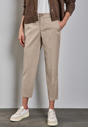 Osoba ubrana w beżowe spodnie 3/4, białe sneakersy z różowymi akcentami, białą koszulę i brązową zapinaną na guziki kurtkę, stojąca z rękami w kieszeniach.