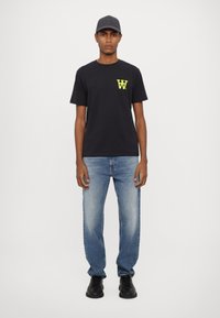 Schwarzes Baumwoll-T-Shirt mit neon-gelbem Logo auf der Brust, kombiniert mit hellblauen Jeans und schwarzen Schuhen. Das Modell steht vor einem weißen Hintergrund.