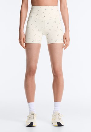 Femme portant un short de vélo crème taille haute avec un petit imprimé floral, des chaussettes blanches montantes et des chaussures de sport crème, debout sur un fond blanc.