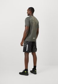 T-shirt vert olive avec un graphique de serpent et du texte au dos, associé à un short noir à motifs avec un dessin de crâne. Chaussures de sport noires.